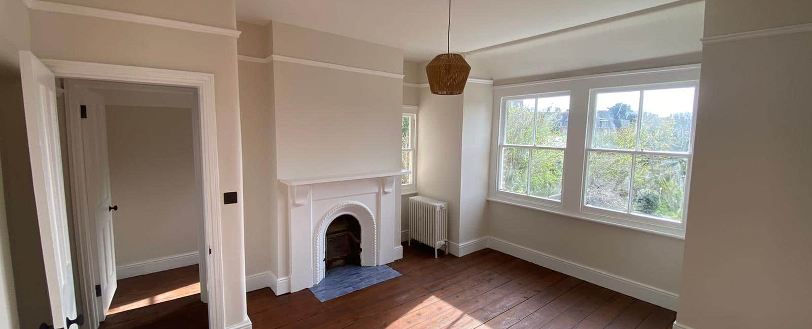 Bright empty room with wooden floors, a fireplace, large windows, and a pendant light in a renovated home setting