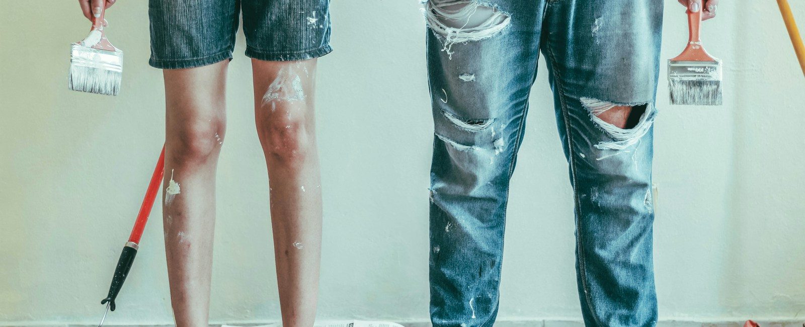 Barefoot couple in denim holding paintbrushes stands on newspaper-covered floor during DIY painting project