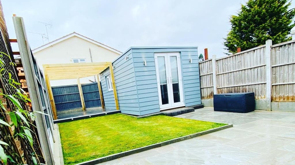 Compact modern garden with wooden pergola, grey garden room, and manicured lawn enclosed by wooden fences on a cloudy day
