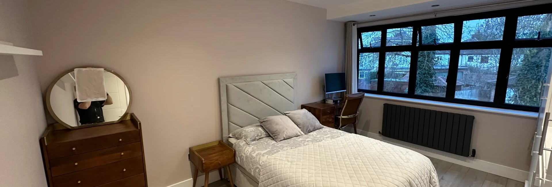 Modern bedroom with grey decor, wooden furniture, and large windows featuring a desk and chair setup for work or study
