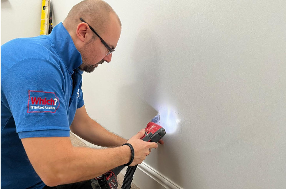 Person in blue shirt using a power tool on a wall indoors, displaying a trusted trader badge on sleeve