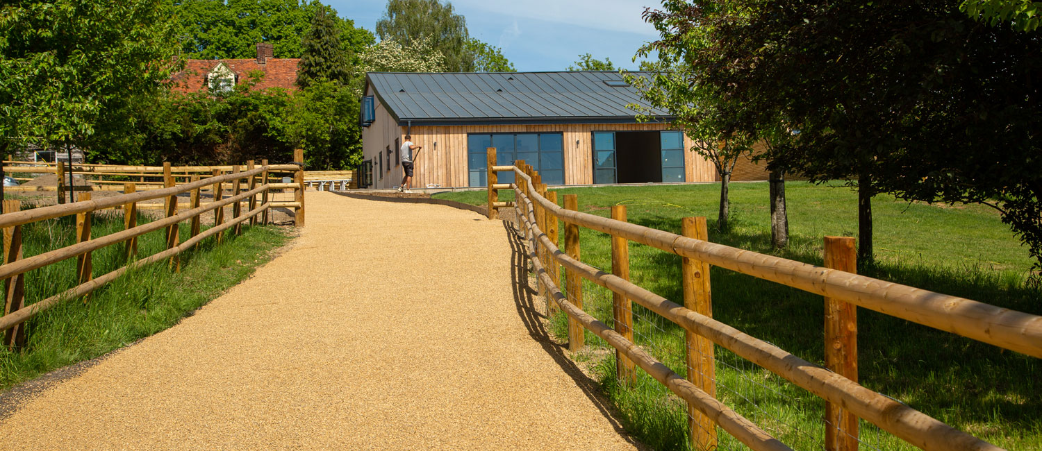 Scenic countryside path with wooden fence leading to modern barn-style house amidst greenery