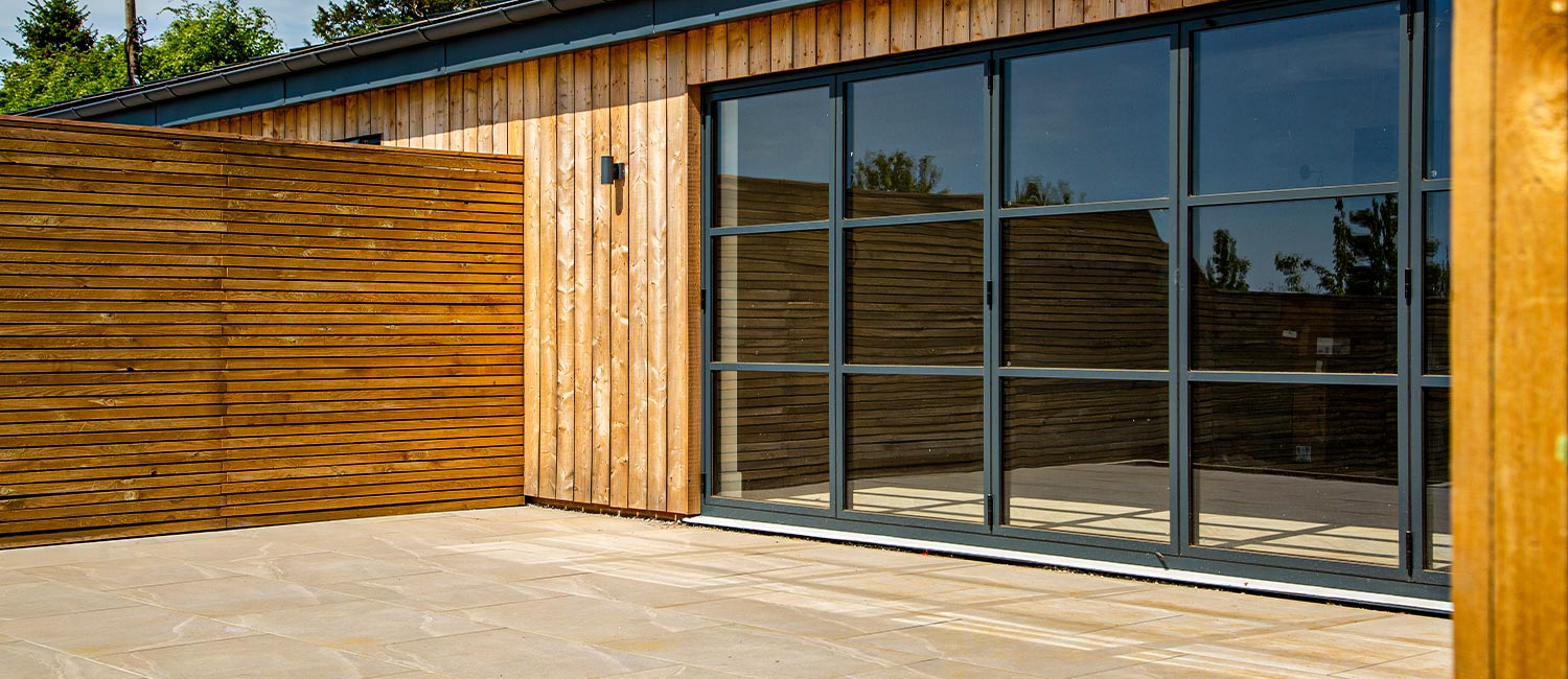 Modern wooden building exterior with large glass doors and spacious stone patio in a sunny garden setting