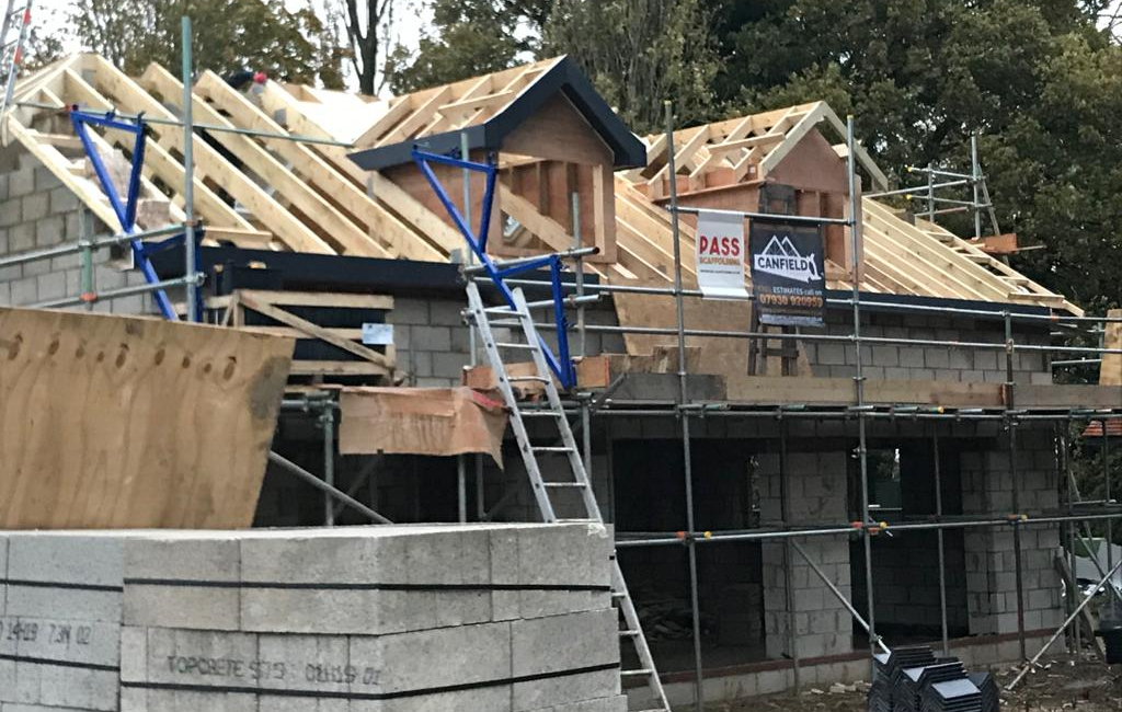 Construction site with partially built house featuring wooden roof trusses and scaffolding, surrounded by trees