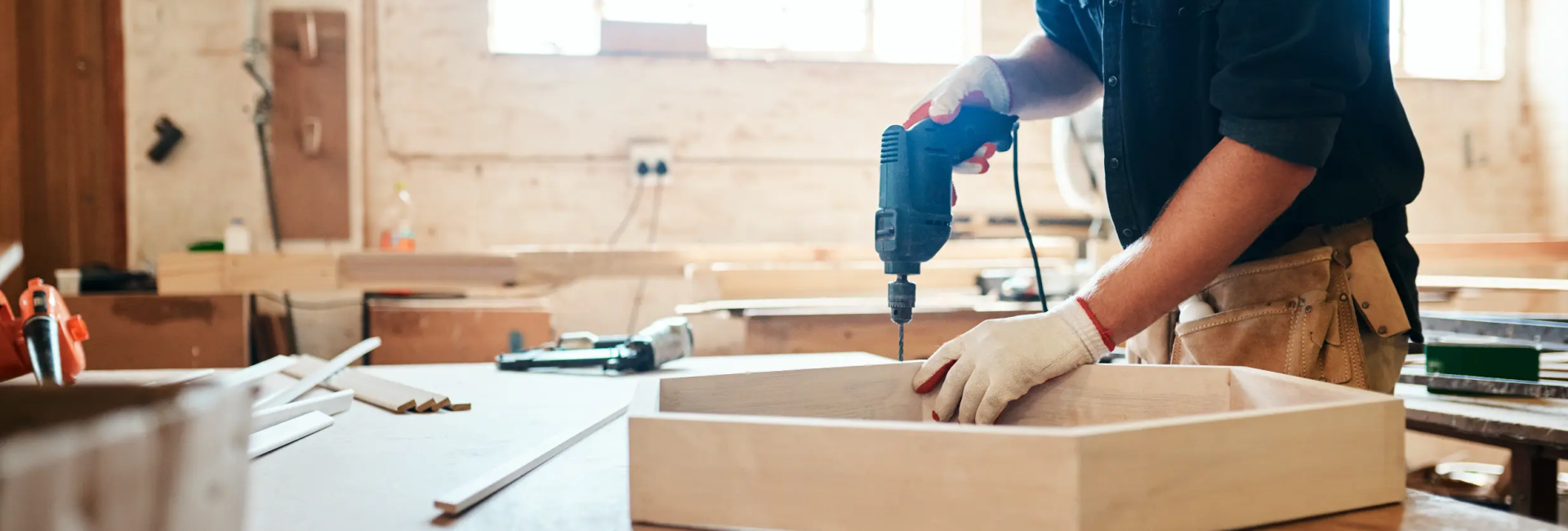 Carpenter using an electric drill on a wooden project in a sunlit workshop, highlighting craftsmanship and skill