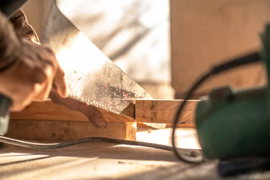 Carpenter using hand saw to cut wooden plank in sunlight, showcasing woodworking skills and craftsmanship
