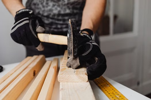 Person wearing gloves hammering nails into wooden planks with a tape measure on the workbench