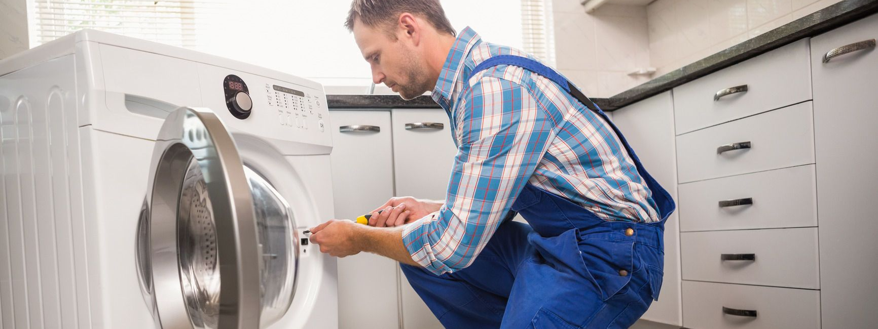 Man repairing a washing machine in a modern kitchen setting wearing blue overalls with a focused expression
