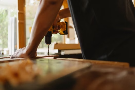 Person using a cordless drill on a wooden plank in a workshop setting