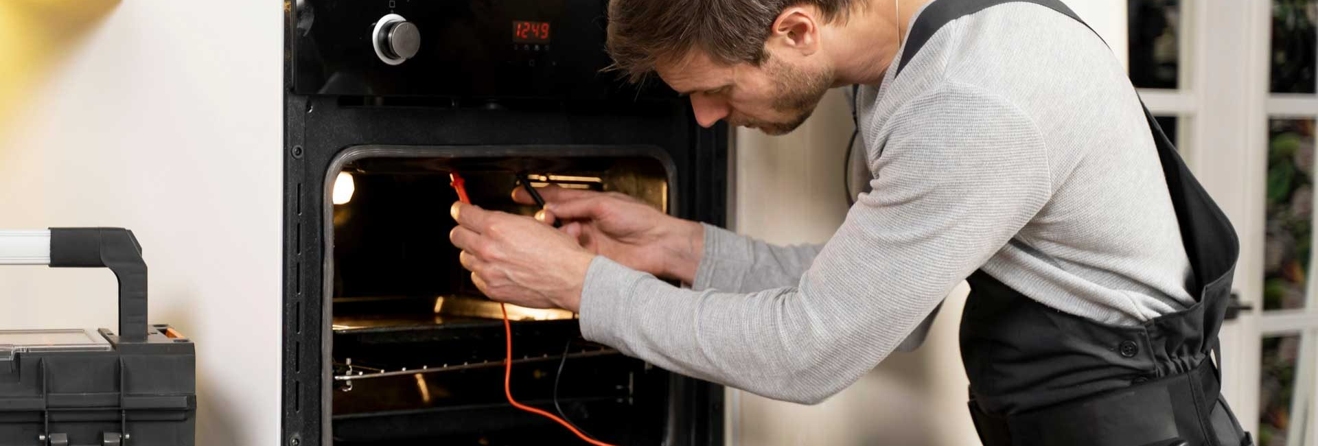 Technician testing oven temperature with multimeter, wearing work apron, tool box nearby in bright kitchen setting