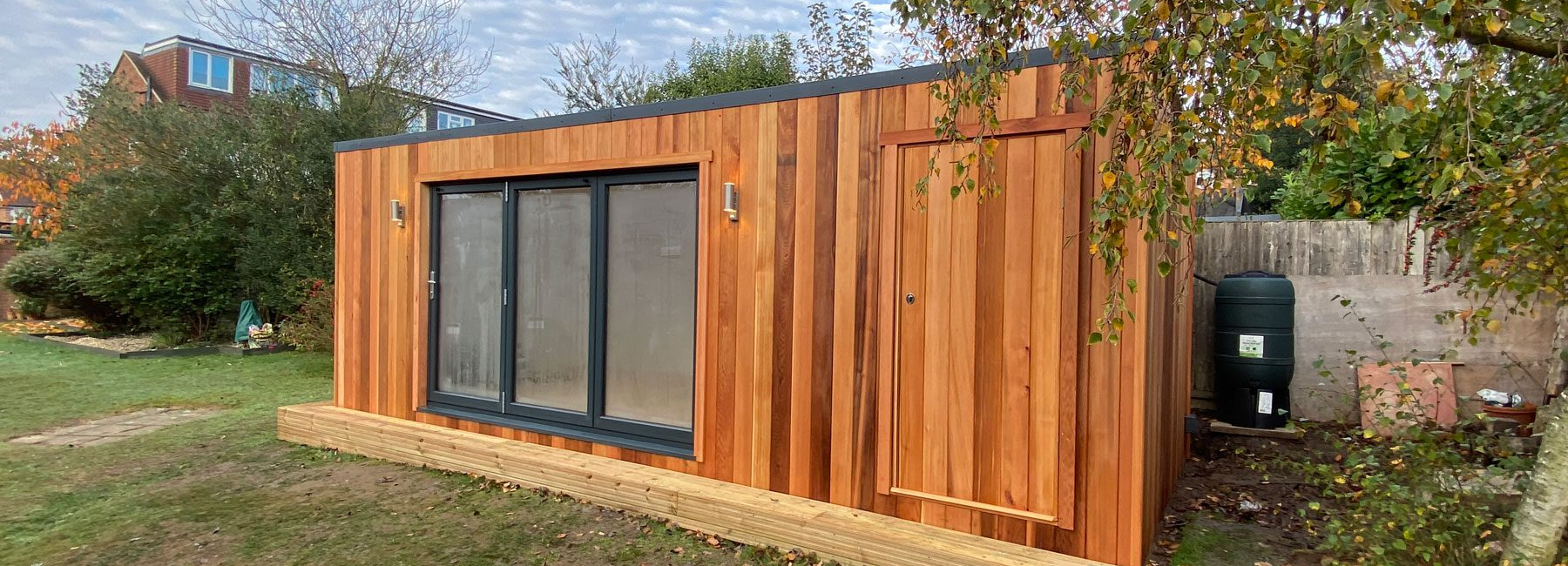 Wooden garden studio with large glass doors in lush backyard setting under a partly cloudy sky
