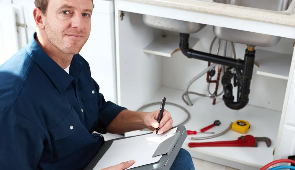Plumber in blue shirt inspecting kitchen sink pipes while taking notes clipboard next to tools and open cabinet