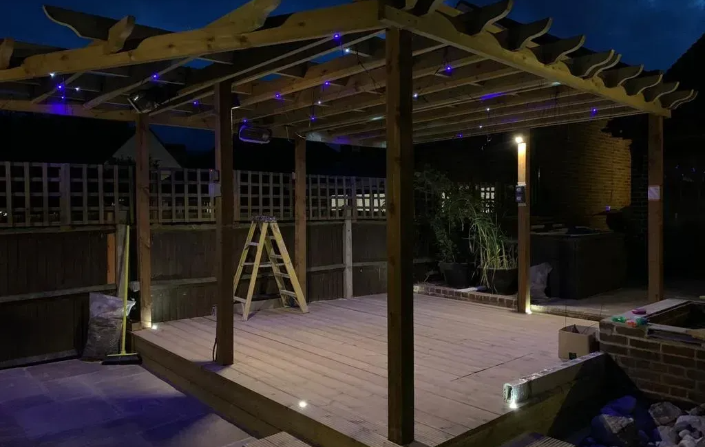 Wooden pergola with night lighting and ladder, set on a patio deck, framed by wooden fencing and plants