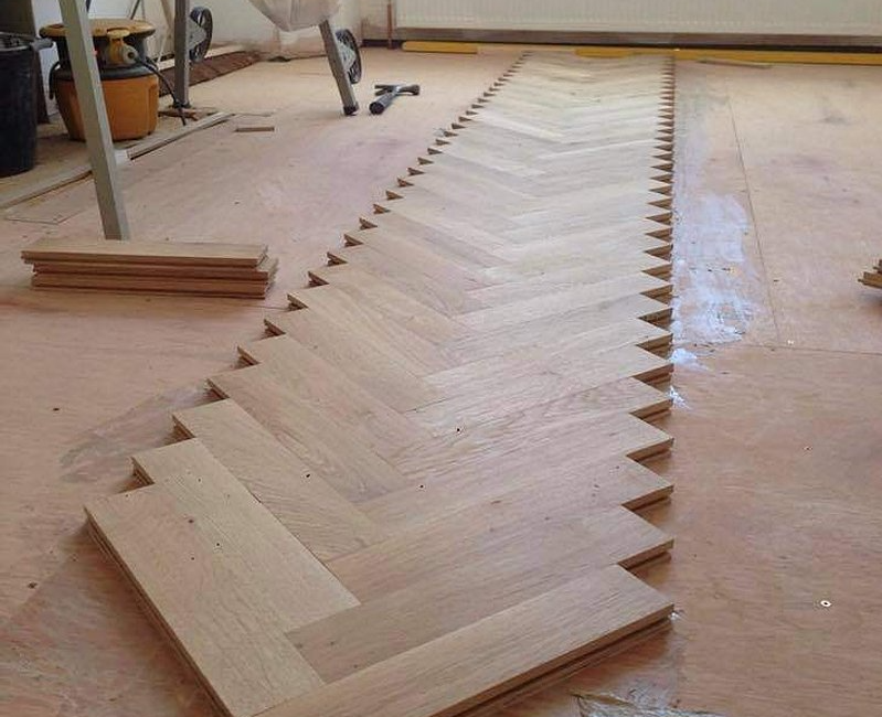 Herringbone parquet flooring installation process in progress at a construction site with tools and planks visible