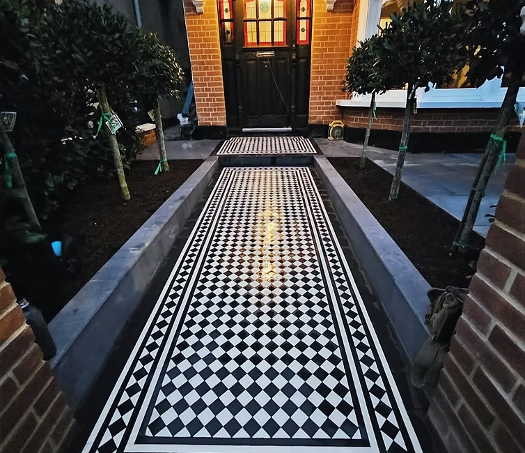 Victorian-style tiled pathway leading to an inviting entrance with black door and decorative windows, flanked by greenery