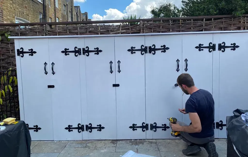 Man installing black hinges on white outdoor wooden storage cabinets