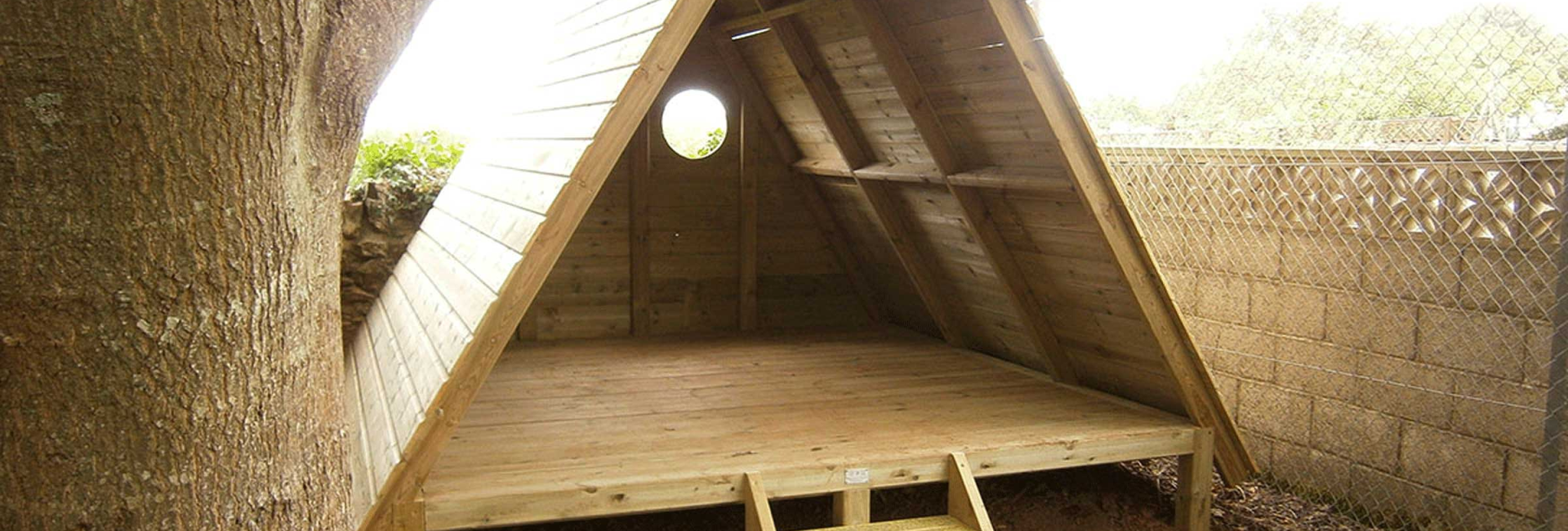 A-frame wooden shelter with ladder under a large tree with leaves and enclosed by a chain-link fence in a garden setting