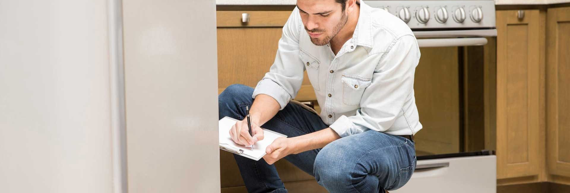 Man inspects kitchen appliance, notes repairs in notebook, toolkit open nearby, wearing jeans and work boots