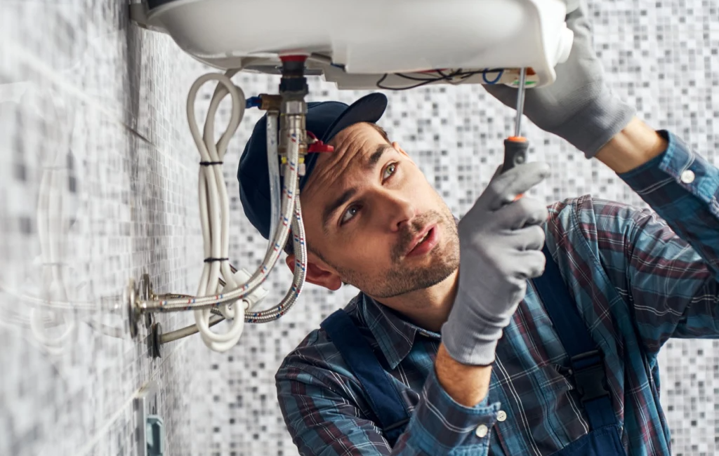 Electrician in plaid shirt and cap repairing a white water heater on a tiled wall using a screwdriver and gloves