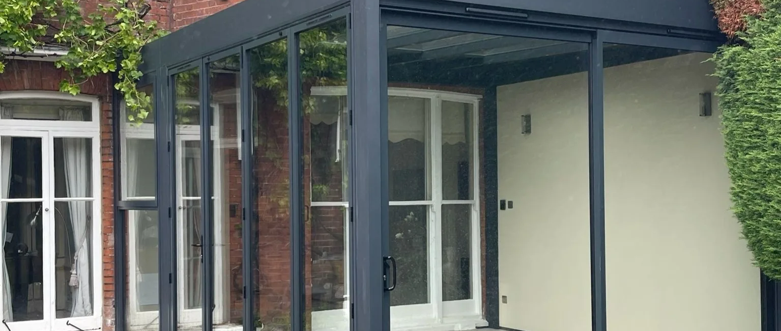 Modern glass conservatory extension on a brick house surrounded by greenery and a stone patio