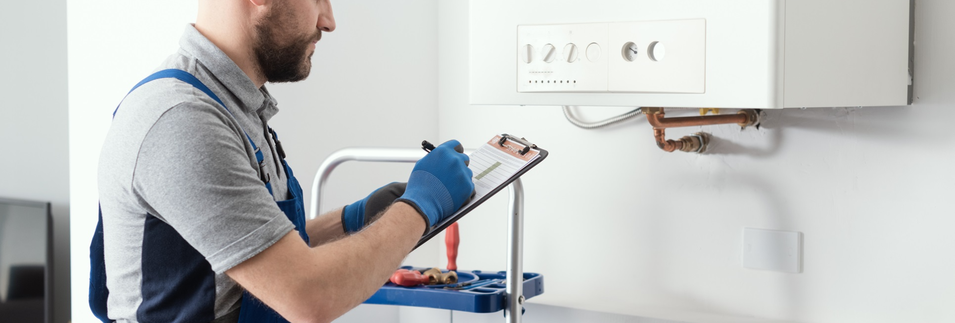 Technician inspecting boiler with clipboard, wearing blue overalls and gloves in a modern home setting