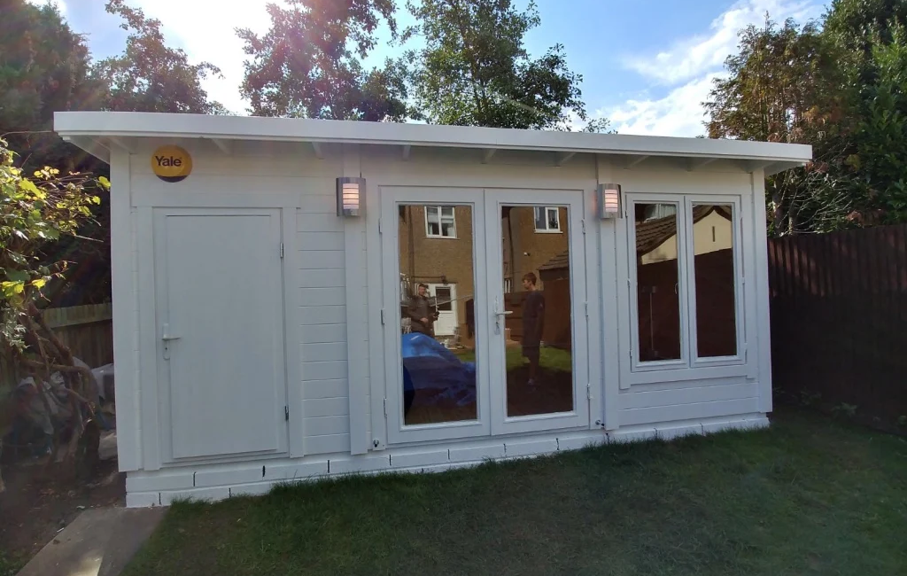 White garden shed with large windows and a Yale lock, surrounded by trees and a wooden fence on a sunny day