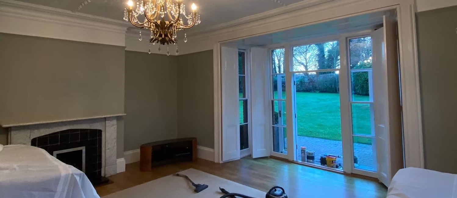 Living room with chandelier, open French doors, fireplace, and vacuum on carpet, overlooking a garden lawn
