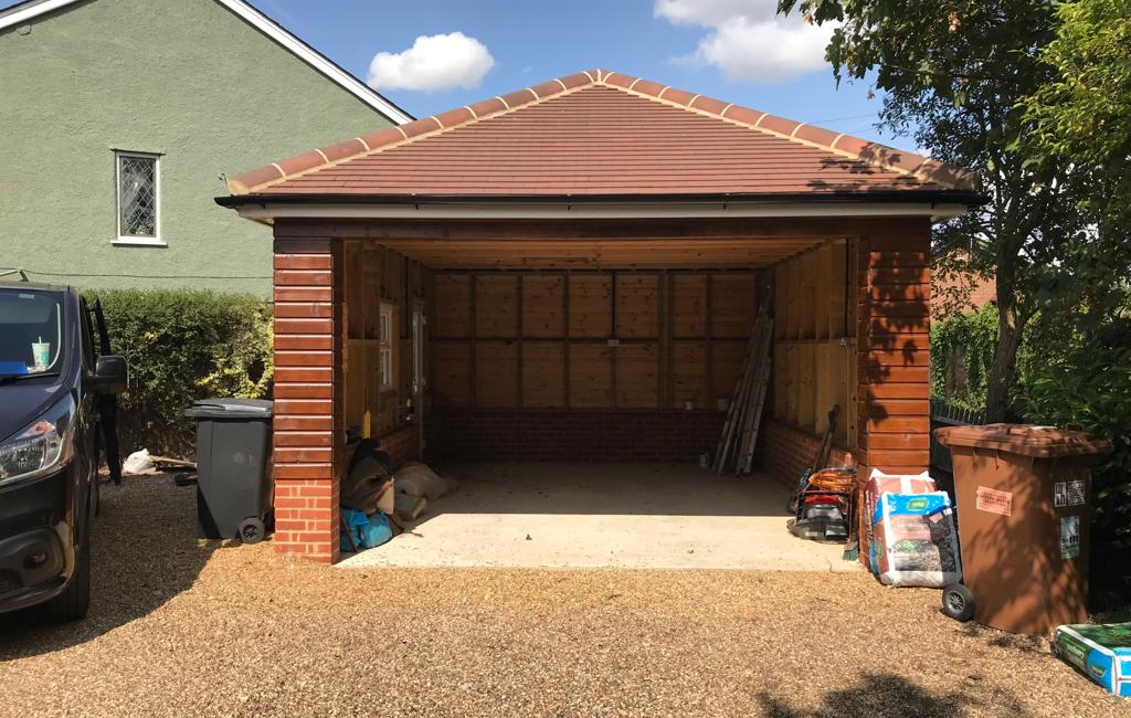 Empty wooden garage with open doors in residential area with parked van, bins, and garden supplies nearby
