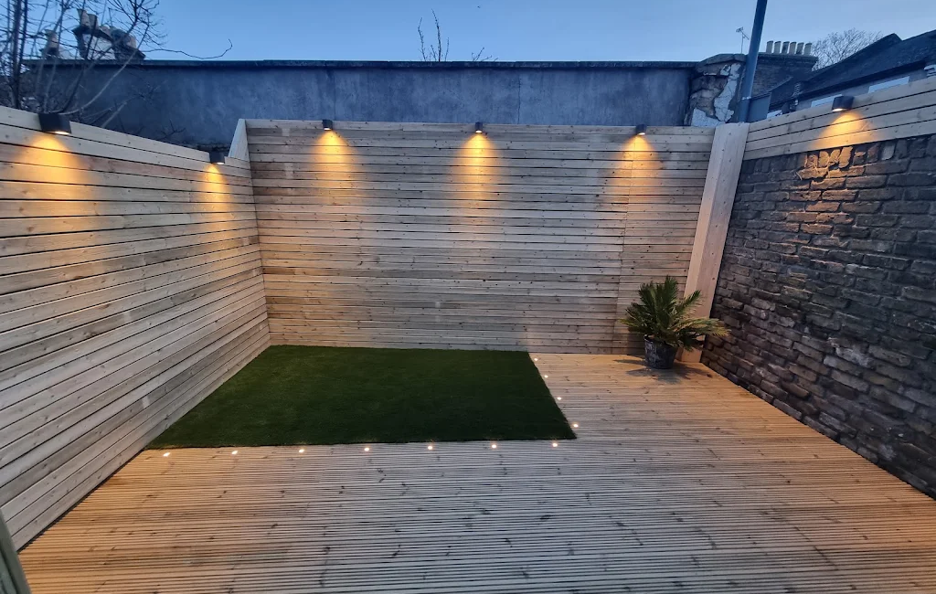 Modern wood-panelled garden with decked flooring and ambient lighting surrounding a small artificial grass area