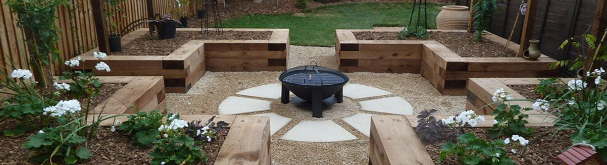 Wooden raised garden beds with a central fire pit, surrounded by greenery and flowers in a backyard setting