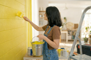 Woman painting a wall bright yellow with a brush, wearing overalls and holding a paint bucket in a sunlit room