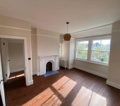Bright empty room with wooden floors, a fireplace, large windows, and a pendant light in a renovated home setting