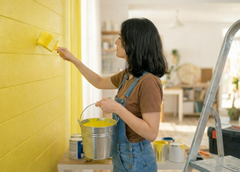 Woman painting a wall bright yellow with a brush, wearing overalls and holding a paint bucket in a sunlit room