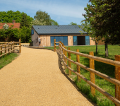 Scenic countryside path with wooden fence leading to modern barn-style house amidst greenery