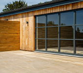 Modern wooden building exterior with large glass doors and spacious stone patio in a sunny garden setting