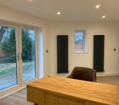 Modern home office with large wooden desk, floor-to-ceiling windows, black radiators, and outside garden view