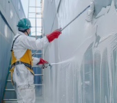 Worker in protective gear painting a large container white with a roller brush outdoors
