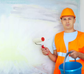 Construction worker holding paint roller and blue bucket, wearing orange high-visibility vest and helmet near painted wall