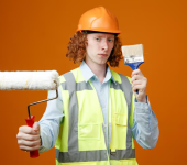 Construction worker holding a paint roller and brush, wearing a hard hat and safety vest against an orange background