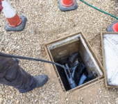 Worker checking underground cables in an open manhole with safety cones on gravel surface