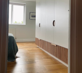 Modern bedroom with wooden flooring and white built-in wardrobes next to a window allowing natural light
