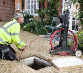 Man in high-vis vest uses reel to feed cable into drain for maintenance work in residential garden setting