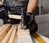 Person wearing gloves hammering nails into wooden planks with a tape measure on the workbench