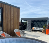 Modern outdoor space with hot tub, wooden garden room, cosy seating area, and vibrant patio plants under a clear sky