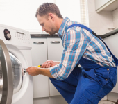 Man repairing a washing machine in a modern kitchen setting wearing blue overalls with a focused expression