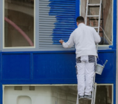Painter on ladder applying blue paint to building exterior for renovation project