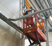 Two technicians in safety gear working on duct installation from a scissor lift inside a large industrial facility