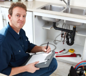 Plumber in blue shirt inspecting kitchen sink pipes while taking notes clipboard next to tools and open cabinet