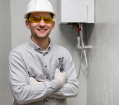 Smiling engineer with hard hat, safety glasses, and tools standing next to a modern boiler in a grey utility room