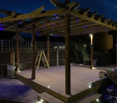 Wooden pergola with night lighting and ladder, set on a patio deck, framed by wooden fencing and plants